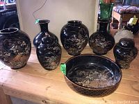Group photo showing seven matching Japanese pottery pieces with black glaze and intricate floral chrysanthemum designs