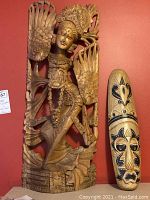 Front view of the hand carved wood sculpture with detailed figure and wings next to the tribal mask on a red background.