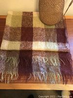 Full view of folded mohair blanket showing checkered pattern in cinnamon, cream, and pale green colors, placed on wooden surface next to a woven vase.