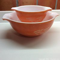 Two nested vintage Pyrex handled bowls in tangerine color with white wheatsheaf print on sides and dual pouring spouts on handles.