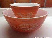 Two nested vintage Pyrex mixing bowls with tangerine exterior and white wheatsheaf pattern around sides, white interiors visible.