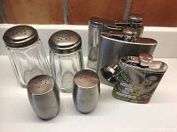 Overview photo showing all seven items including two glass sprinklers, two stainless steel salt and pepper shakers, and three stainless steel flasks (one with graphic).