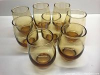 Seven amber-colored glass tumblers arranged in a group on a white surface showing their size and color.