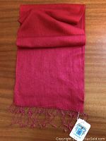 Full view of folded cranberry pashmina scarf showing fringed ends with tag on wooden surface.