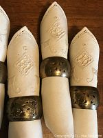 Close-up showing white linen napkins folded inside brass napkin rings, highlighting embroidered and cut-out details on napkins and etched designs on brass rings.