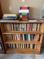Bookshelf with multiple paperback and hardcover books organized on three shelves and additional stacked books on top.