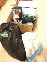 Photo showing several camping items including a cardboard box with a white resin cooler on top, a green tent or tent fly inside the box, air mattress or bag, and a rolled-up blanket next to the box and black bag labeled 'sleeping bag'.