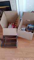 Photo showing two cardboard boxes containing VHS tapes, DVDs, and a Scrabble game box, along with a wooden basket on the floor.
