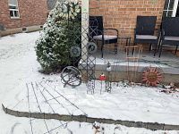 Full view of the garden trellises, sun ornament, and wire cages partly covered in snow outside a brick house patio with chairs.