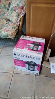 Original boxed Instant Pot Viva 8 quart multi-use pressure cooker next to floral couch and wooden furniture.