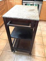 Wood veneer kitchen cart with granite-style stone top, wood drawer with knob, two open shelves, caster wheels, and side handle. Shows visible wear and slight wobble.