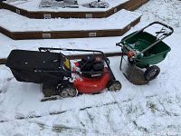 Side view of red Yard Machines lawn mower with attached grass catcher and green Scotts seed spreader behind it on snow-covered ground.