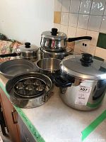 Full set of stainless steel pots and pans displayed together showing multiple sizes of pots with lids and black knobs/handles along with a round baking pan and springform pan.