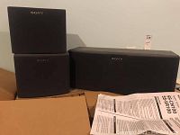 Three Sony speakers stacked; one larger center speaker and two smaller side speakers, all black with Sony logo and in original boxes.