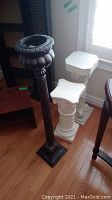 Set of three composite plant stands including two white decorative columns and one taller brown pedestal on hardwood floor near window.