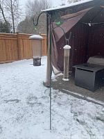 Bird feeders mounted on a metal stand in a snowy backyard with patio furniture and propane tank visible in background.