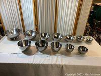 Full set of 12 stainless steel mixing bowls arranged in two rows on a white tablecloth, showcasing size variation from smallest to largest.