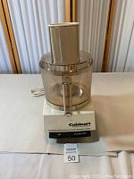 Front view of Cuisinart DLC-7E food processor showing motor base with control buttons and transparent plastic bowl with feed chute.