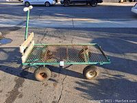 Side angled view of flat bed wagon showing green metal frame, mesh steel bed, wooden handle, and rubber wheels with rust on axles.
