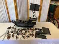 Large pirate ship with black hull and mast, fabric sails laid out on table, surrounded by multiple pirate figures and accessories