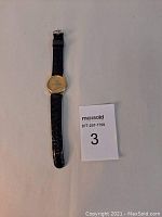 Full view of Seiko Mickey Mouse gold tone watch with black leather strap laid flat on a white surface alongside a Maxsold number card.