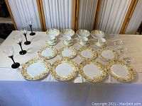 Overall photo of full set showing 4 dinner plates, 4 smaller plates, 4 smallest plates, 4 cups with saucers, 1 bowl, 4 black stemmed goblets, and 4 clear lowball glasses on table.