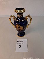Front angle view of a cobalt blue porcelain vase with gold handles and gold painted scene of a couple.