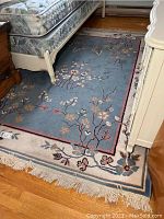 Full view of rectangular floral patterned area rug placed between bed and dresser, showing the blue background with floral designs and beige border, along with slip pad underneath.