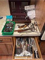 Kitchen counter with boxed Oneidacraft Deluxe stainless flatware, assorted spoons and forks in trays, and serving utensils spread around.