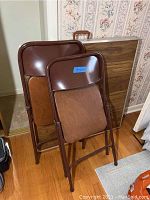 Photo showing two brown metal folding chairs with faux wood seats and backs folded and leaning against a wall, next to a vertical board likely the folded table top.