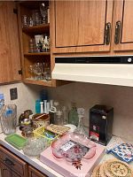 Photo shows assorted glassware and kitchen items on a countertop, including glass decanter, potpourri crock, glass canisters, pink serving dish, towel, and various small kitchen items.