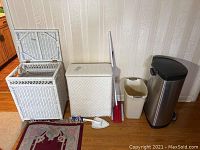 Photo shows the entire lot including a white wicker hamper with hinged lid open, a white plastic wicker-look hamper, a stainless steel SimpleHuman trash can, small white waste bin, and cleaning tools (broom, duster, dustpan) arranged against a wall.