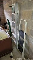 Photo showing the Keller 6-foot aluminum ladder, the white 3-step folding step stool, and green convertible hand truck leaning against a concrete wall with visible scratches and use marks.