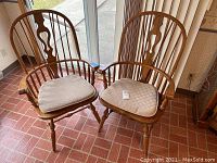 Two oak colonial style dining arm chairs with cushioned seat pads shown near window, displaying overall condition and design details.