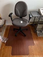 Full view of gray fabric office chair with black base and armrests on a clear plastic floor mat on hardwood floor.