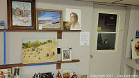 Wide shot showing various paintings on shelves and walls including a snowy barn scene, beach, portrait and autumn landscape