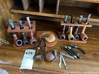 Overview of nine pipes arranged in two wooden pipe stands, one separate pipe bowl and four stems, along with pipe cleaner tools on a wooden surface.