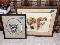 Two framed and matted prints leaning against a couch; one print depicts two dogs, the other a raccoon.