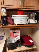 Photo showing multiple kitchen appliances on wooden shelves including Hamilton Beach slow cooker, food processor, George Foreman grill, Dazey Chefs Pot, and frying pan