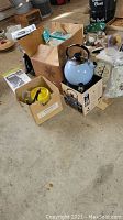Photo showing multiple items including blue and yellow tea kettles, boxed tea kettle and coffee maker on floor.