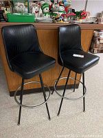 Two matching black vinyl bar stools with metal legs and circular footrests seen side by side near a wooden cabinet.