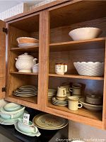Wood cabinet open showing multiple stoneware dishes, mugs, bowls, and milk glass bowl on shelves