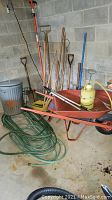 Red metal wheelbarrow, assorted yard tools, green garden hose, and rusted metal trash can in concrete corner
