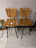 Two matching wood seat stools with metal frames shown side by side, in a basement setting, carpeted floor and paneled wall in background.