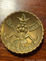 Top view of antique brass pin dish with devil or pixie figure, 'LUCKY PIXIE' text, scalloped edges.