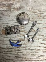 Top view of antique sterling silver lidded container, two silver spoons, a decorative buckle, and an enamel bird brooch laid on wooden surface