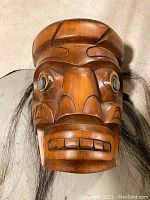 Front view of carved cedar mask showing facial features, shell inlay eyes, and attached hair.