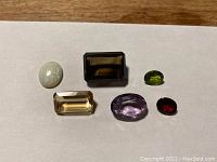 Six loose gemstones arranged on white background for identification and sale. Includes various cuts and colors corresponding to amethyst, smoky quartz, opal, citrine, garnet, and peridot.