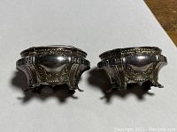 Front and side view of both silver salt cellars side by side displaying design details and feet.
