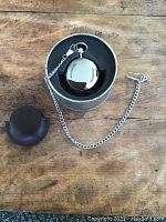 Round metal tin case with silver-tone pocket watch and leather pouch on wood surface